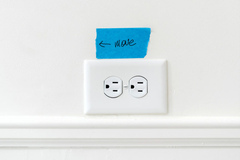 Close up of electrical outlet with piece of blue painter's tape above it that has "Move" written on it