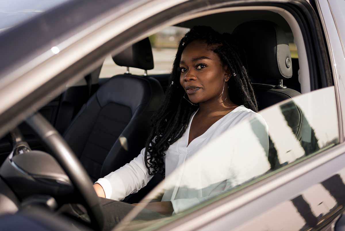 Millennial-age woman seated in driver’s seat of a car, looking out the open window at the camera.