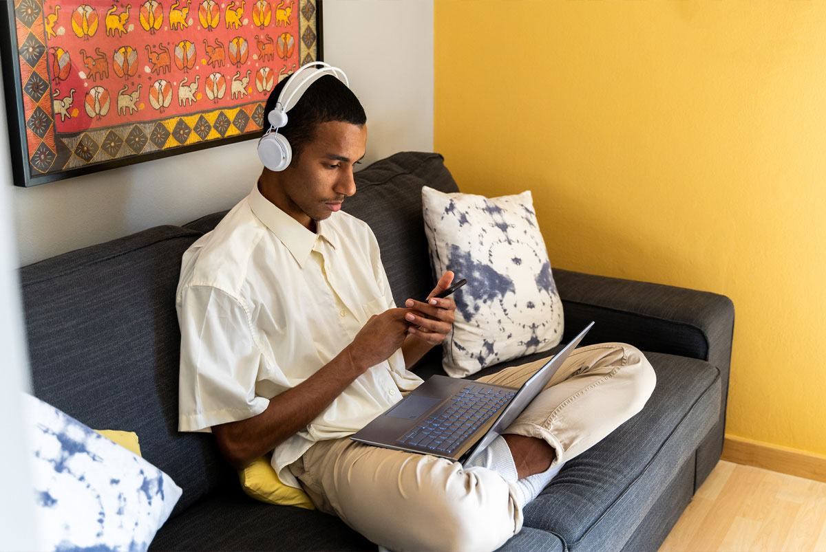 Young man sitting on couch with headphones on with his laptop on his lap and looking at his phone