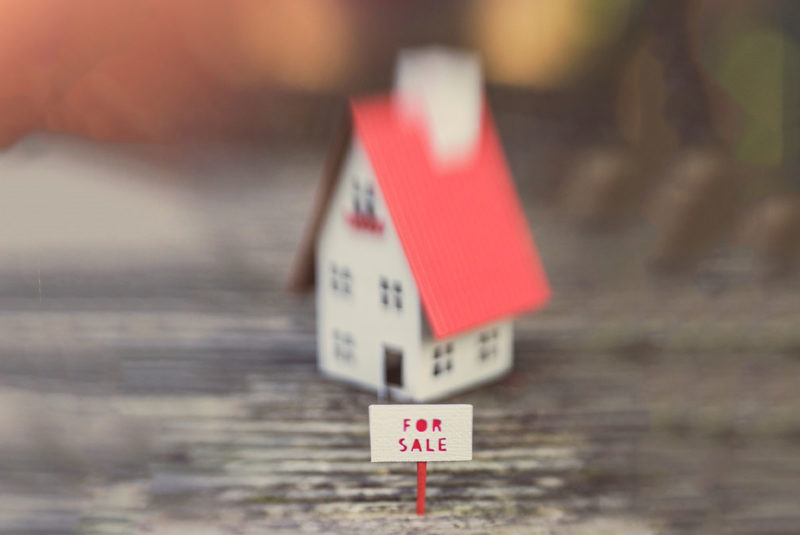 A tiny little cream and red paper house with a "for sale" outside it