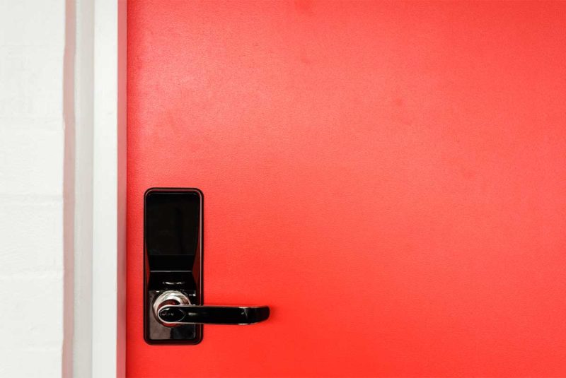 Closeup of a red door with a black handle and card reader, much like you’d find at a hotel.